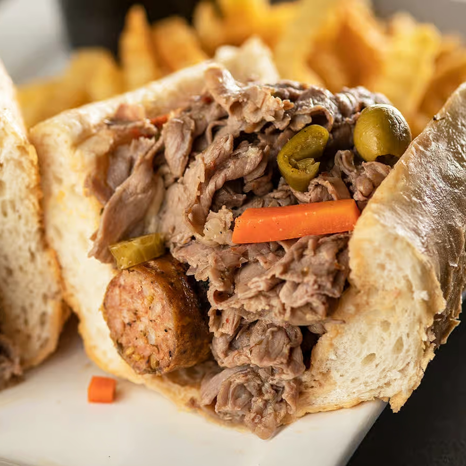 Close-up of a Papa Saverio's Italian beef sandwich with sausage, peppers, and carrots, with fries in the background.